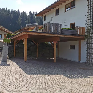 Ein modernes Haus mit einer überdachten Terrasse und einem gepflasterten Parkplatz. Im Hintergrund sind grüne Bäume und eine ruhige Landschaft sichtbar.