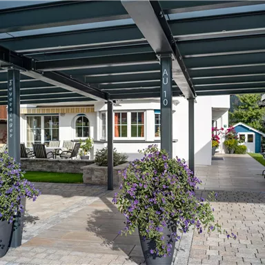 A covered terrace with modern furniture and blooming plants. In the background, a house and a small garden are visible.