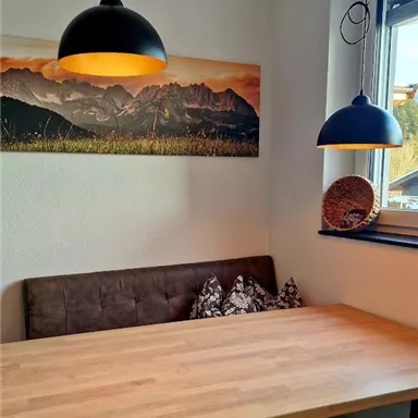 Ein gemütlicher Essbereich mit einem Holz-Tisch und einer Couch. Über dem Tisch hängen zwei stilvolle Lampen und an der Wand ist ein Bild von Bergen.
