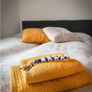 A cozy bed with orange pillows and fresh, yellow towels. Next to the towel is a colorful keychain.