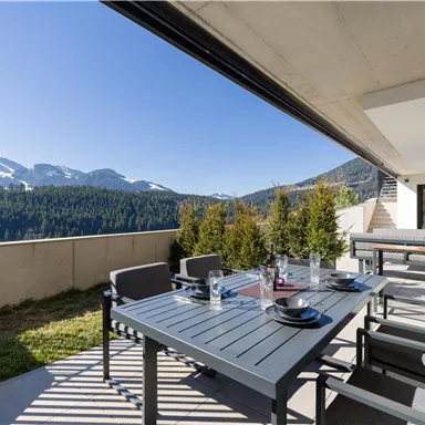 Eine moderne Terrasse mit einem Esstisch und Stühlen, umgeben von einer beeindruckenden Berglandschaft. Im Hintergrund sind schneebedeckte Gipfel und ein klarer blauer Himmel sichtbar.