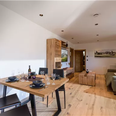 Ein modernes Wohnzimmer mit einem Esstisch, Stühlen und einer gemütlichen Couch. Die hellen Farben und Holzmöbel schaffen eine einladende Atmosphäre.