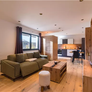 Ein modernes Wohnzimmer mit einem bequemen Sofa und einem Holzstamm-Tisch. Die offene Küche und große Fenster sorgen für einen hellen, einladenden Raum.