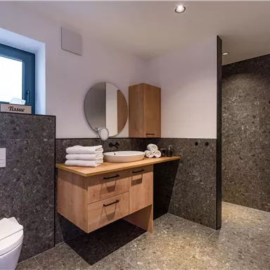 Ein modernes Badezimmer mit grauen Wänden aus Stein und einem eleganten Holzwaschbecken. Es gibt große Fenster, die viel Licht hereinlassen.