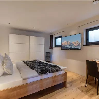 Ein modernes Schlafzimmer mit einem großen Bett und neutraler Einrichtung. Ein Fernseher und ein Schreibtisch ergänzen den Raum, der gut beleuchtet ist.