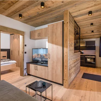 Ein modernes Wohnzimmer mit Holzwänden und einem Fernseher. Die offene Küche ist gut ausgestattet und der Raum wirkt gemütlich.