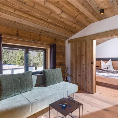 Ein gemütliches Zimmer mit Holzwänden und einem hellen Sofa. Im Hintergrund sieht man ein Bett und große Fenster mit Blick auf die Natur.