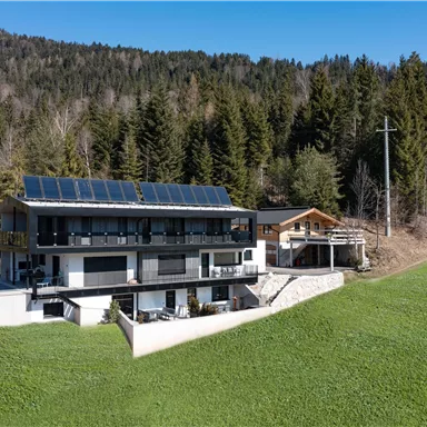 Ein modernes Haus mit Solarpanelen, umgeben von einer grünen Wiese und Wald. Im Hintergrund sind weitere Gebäude und ein klarer blauer Himmel zu sehen.