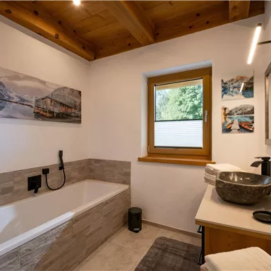 A modern bathroom with a bathtub, wooden ceiling, and large windows. The walls are decorated with stylish pictures and the fittings are elegant and functional.