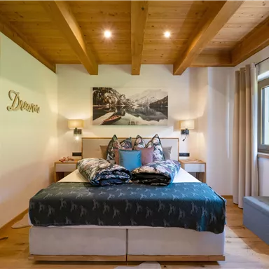 A cozy bedroom with a wood ceiling and a large bed. A picture of a mountain landscape hangs on the wall, and the window offers a view of greenery.