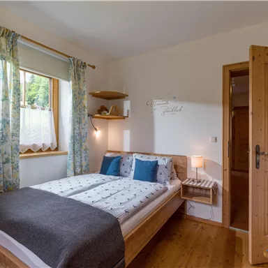 A cozy bedroom with a double bed and blue pillows. The walls are light, and daylight streams in through the window.