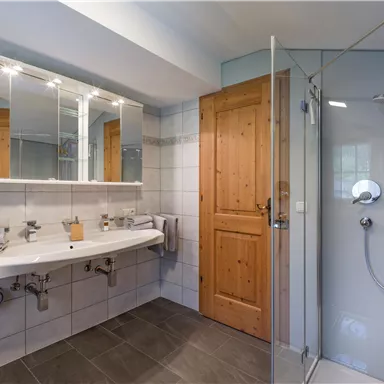 A modern bathroom with a shower, a double washbasin, and large mirrors. The room has bright tiles and a wooden house door.