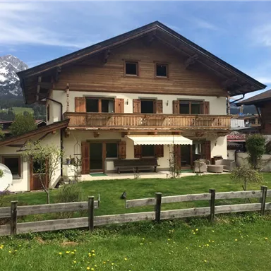 Ein schönes Holzhaus mit Balkon und Garten. Im Hintergrund sind die Berge zu sehen.