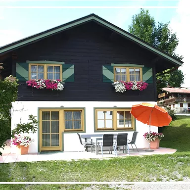A charming two-story wooden house with colorful flower windows. In front of the house, there is a table with chairs and a red umbrella.