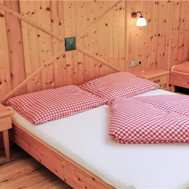 A cozy bedroom with a wooden bed and checkered cushions. The walls are paneled with wood, and there is a comfortable seating area in the background.