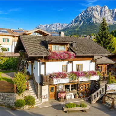 Ein malerisches alpenländisches Haus mit bunten Blumenbalkonen, umgeben von grünen Wiesen und markanten Bergen im Hintergrund. Die Sonne scheint und der Himmel ist klar.