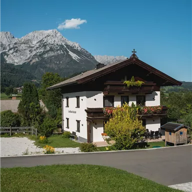 A traditional chalet with a brown roof and colorful flowers. In the background, majestic mountains and a clear blue sky can be seen.