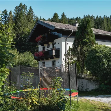 A beautiful house surrounded by trees and plants. In the foreground, there is a trampoline that invites play.