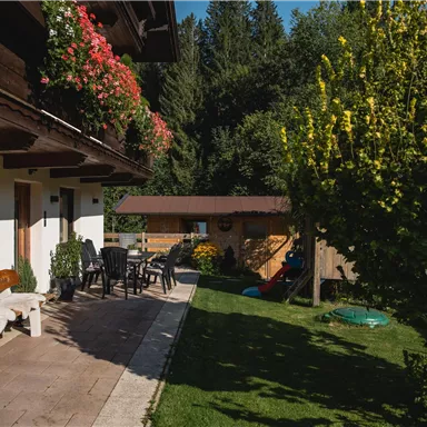 A cozy outdoor area with a terrace, table, and chairs. In the background, trees and a wooden house are visible.