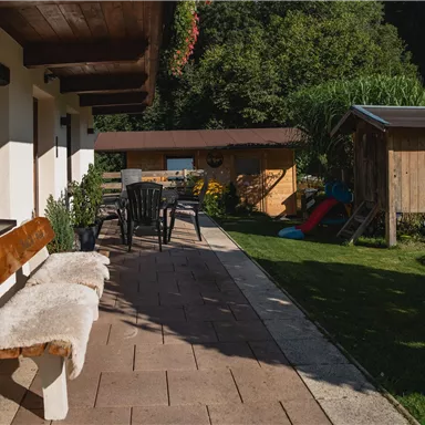 An inviting terrace with a bench and chairs. In the background, there is a garden shed and play equipment on the lawn.