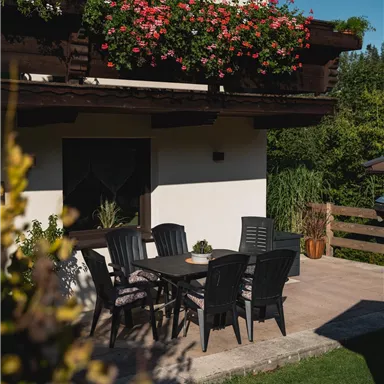 A beautiful outdoor area with a plastic table and chairs. Flowers are hanging from the balcony and the lawn is green and well-kept.