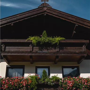 A traditional alpine house with wooden balconies and colorful flowers. Above it rises a cross on the roof under a clear blue sky.