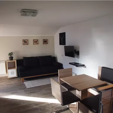 A modern living room with a black sofa and a wooden dining area. Pictures are hanging on the wall, and there is a television.