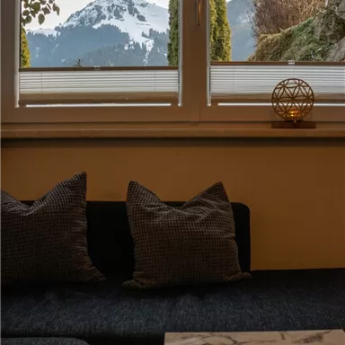 A cozy living room with a view of snow-covered mountains. There are two cushions on the sofa and a small table stands beside it.