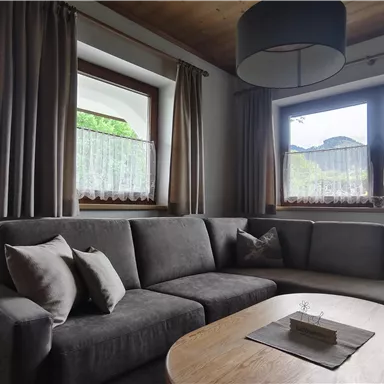 A cozy living room with a gray sofa and a wooden table. The windows are adorned with curtains and provide a view of the outdoors.