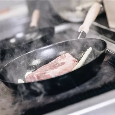 A piece of meat is frying in a pan on the stove. Steam is rising, and kitchen utensils are visible in the background.