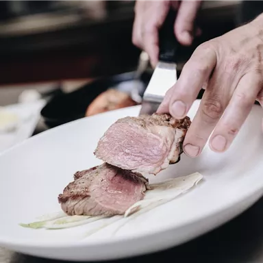 A chef is slicing a piece of juicy meat on a white plate. The dish is lovingly arranged and features fresh side dishes.