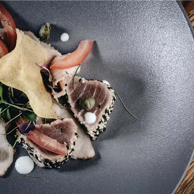 An artfully arranged plate with thinly sliced fish, garnished with fresh herbs and vegetables. The presentation is on a dark, round plate.