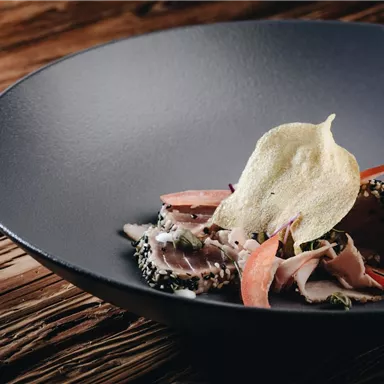 A beautifully arranged dish on a black plate. It contains tender pieces of meat, vegetables, and a crispy decoration.