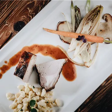 An elegantly arranged plate with grilled meat, vegetables, and a sauce. Served with a side of small dough pieces.
