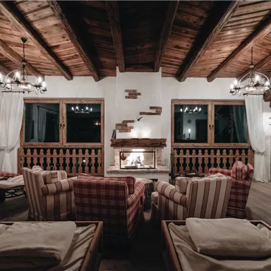 A cozy living space with wooden ceilings and an open fireplace. Comfortable armchairs and windows create an inviting atmosphere.