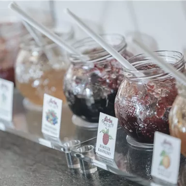 Colorful jars with various jams are standing on a counter. Each jar has a spoon and a small sign with the flavor.