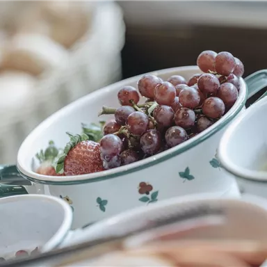 A beautiful fruit platter with grapes and strawberries in a bowl. In the background, more fruits and rolls are visible.