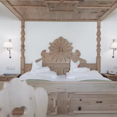 An elegant bedroom with a large, carved wooden bed and two nightstands. The walls are light and the lighting is gentle.