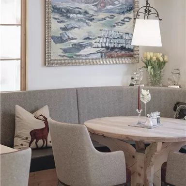 A cozy dining area with a round wooden table and upholstered chairs. A painting of a landscape hangs on the wall.