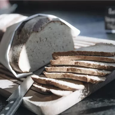 A freshly baked bread lies on a wooden board, partially sliced. Next to it is a knife, and in the background, a sign is visible.
