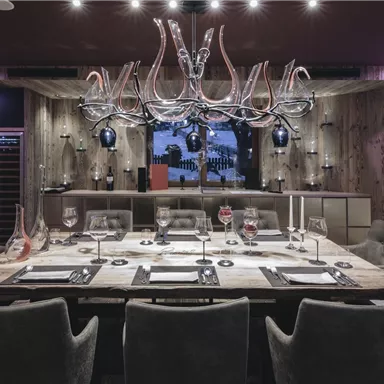 A stylishly furnished dining room with a large dining table and comfortable chairs. In the background, modern light fixtures and a wall with wine bottles can be seen.
