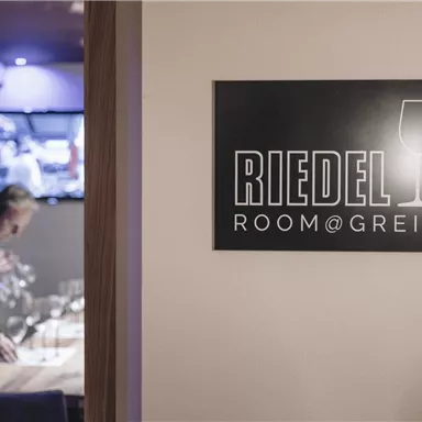 A sign with the inscription "RIEDEL ROOM@GREIL" in an elegant restaurant. In the background, a person is setting up a table.