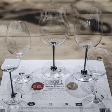 Five wine glasses are standing on a wooden table. Beneath the glasses lies an information sheet with various logos and numbers.