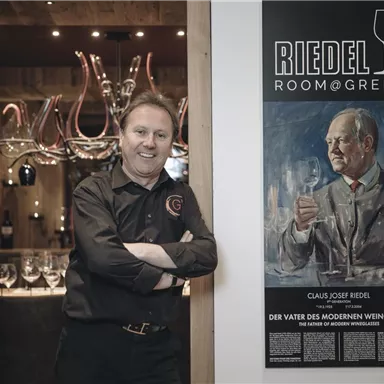A man stands against a wall that displays a portrait of Claus Josef Riedel, the inventor of the modern wine glass. In the background, glasses and wine accessories can be seen.