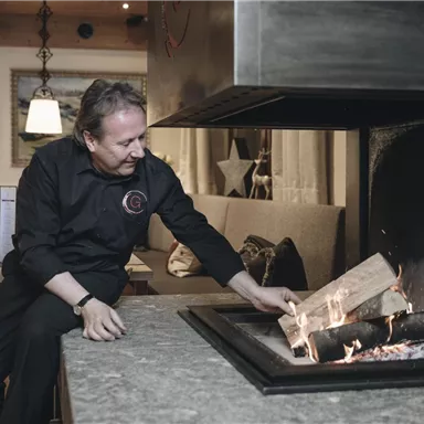 A cook feeds the fire in a cozy restaurant. In the background, comfortable seating and a pleasant atmosphere can be seen.