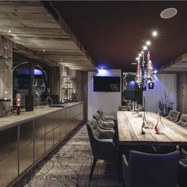An elegant dining room with a long wooden dining table and comfortable chairs. The walls are made of wood, and there is stylish lighting.