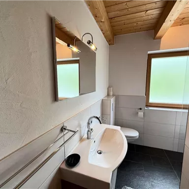 Ein modernes Badezimmer mit Holzdecken und einem großen Spiegel. Die Wände sind hell und der Boden ist dunkel gefliest.
