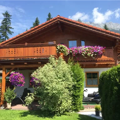 Ein schönes Holzhaus mit einem Balkon, geschmückt mit bunten Blumen. Im Vordergrund gibt es gepflegte grüne Sträucher und Bäume.
