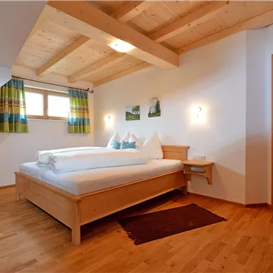 Ein gemütliches Schlafzimmer mit einem großen Bett aus Holz und einem Fenster mit bunten Vorhängen. Der Fußboden ist aus Holz und es gibt einen Stuhl sowie Zugang zu einem Badezimmer.