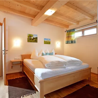 Ein gemütliches Schlafzimmer mit einem Holzbett und zwei Kissen. Die Wände sind hell, und es gibt einige Bilder sowie ein Fenster mit Vorhängen.